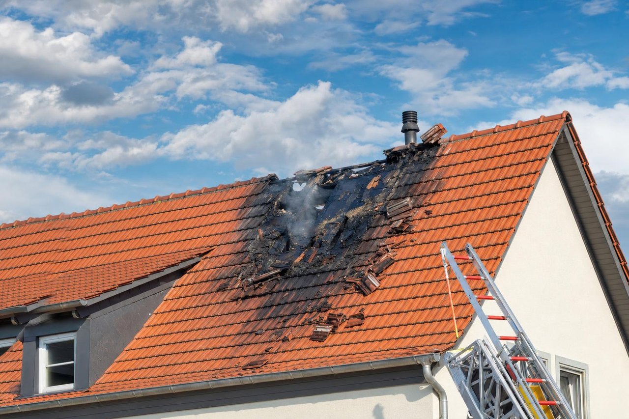 Durch Blitzeinschlag beschädigtes Ziegeldach mit Brandspuren, daneben eine Feuerwehrleiter (Symbolbild)